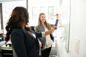 Mujeres escribiendo en pizarra. Lo que alude a que están trabajando en equipo, como lo apunta la gestión colaborativa.