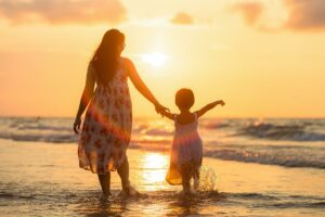 Imagen de una mujer adulta con una niña caminando por la orilla de una playa. La imagen pretende reflejar una relación de madre e hija, para aludir al Día de las Madres.